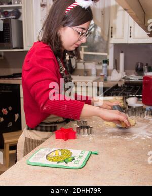 Young girl rolling dough with glasses on counter making cookies in the kitchen with a red sweater on and a santa hat bandana with cookie cutters. Stock Photo