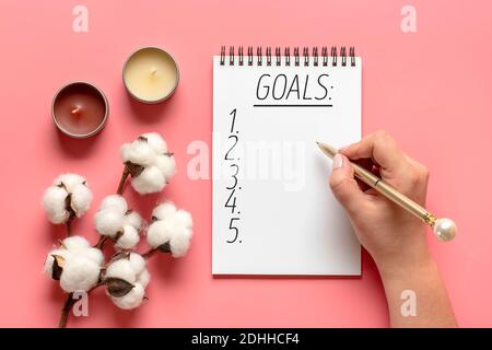 female hand holds pen and writes New Year 2021 goals on white notepad, cactus, candle on pink background Top view Flat lay Business motivation, idea, Stock Photo