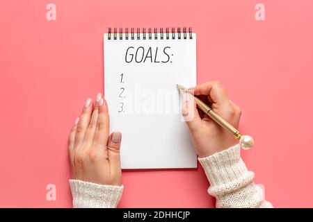 female hand holds pen and writes New Year 2021 goals on white notepad, cactus, candle on pink background Top view Flat lay Business motivation, idea, Stock Photo