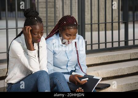 group of young students working together, are sitting on stairs outside with document and a smart phone in casual outfit. Stock Photo