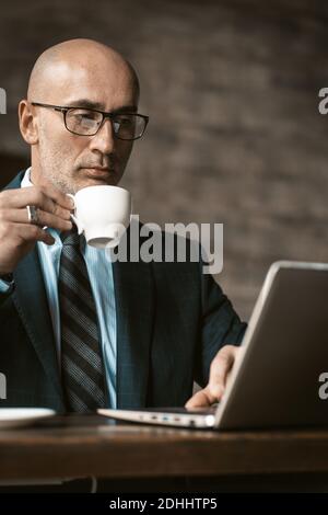 Drinking coffee mature business man working on laptop holding a cup of coffee sitting at table looking at laptop wearing eye glasses. Business concept Stock Photo