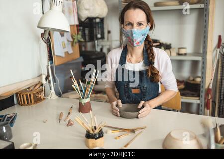 Caucasian woman making ceramic soup plate while wearing protective face mask for coronavirus prevention - Female person at work inside her creative po Stock Photo