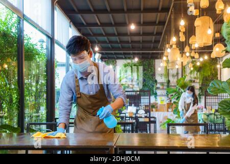 Portrait attractive Asian restaurant employee wear face mask and face shield cleaning table and floor with alcohol and wet wipe before welcome custome Stock Photo