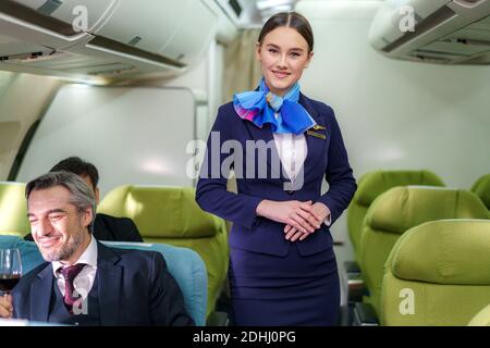 Portrait young woman in blue suit, flight attendant/air hostess in business class cabin at airplane. Stock Photo