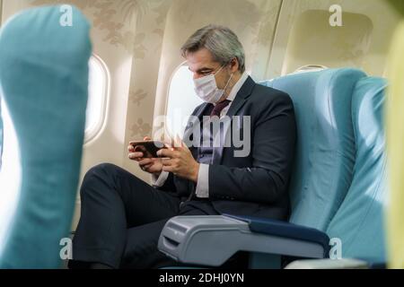 Man wearing face mask and using phone inside airplane during flight. Themes new normal, coronavirus and personal protection. Stock Photo