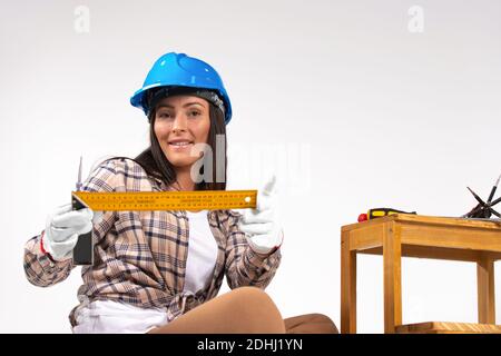 A woman builder with a smile holds a level in her hands next to a stool and tools on a white background. Stock Photo