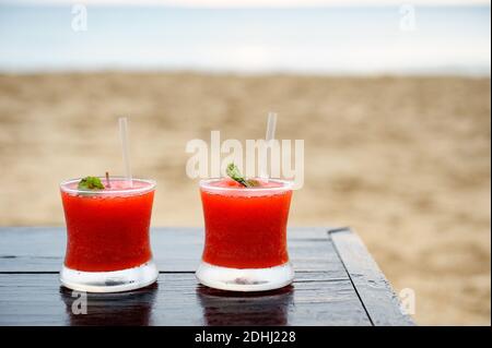Two red cocktails frozen glasses on wet wooden table at beach bar restaurant. View at the ocean and overcast sky. Stock Photo
