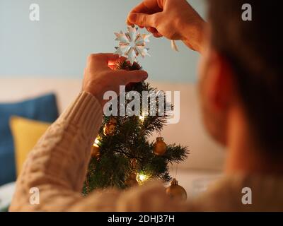 Caucasian young man next to a Christmas tree in a woollen sweater holding a present in his hands. He enjoy the holidays in a cozy home. Stock Photo