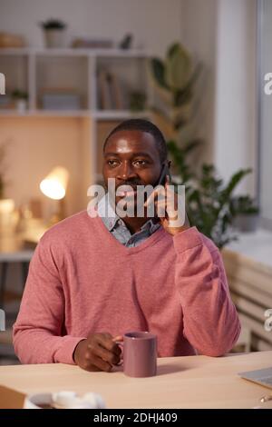 African young man talking on mobile phone while sitting at the table at office drinking coffee Stock Photo
