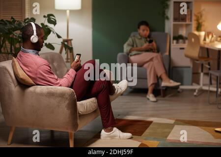 African man in wireless headphones sitting on sofa and using his mobile phone to listen to music with woman using her phone in the background Stock Photo