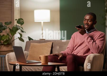 African businessman sitting on sofa in front of table with laptop and recording audio message on his mobile phone he working at home Stock Photo