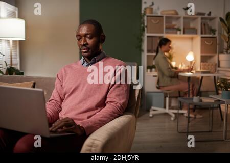 African man sitting on sofa typing on laptop with his colleague working at her workplace in the background Stock Photo
