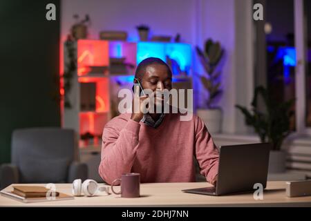 African young businessman sitting at his workplace typing on laptop and has a conversation on the phone working till evening at office Stock Photo