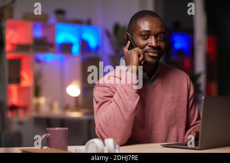 Portrait of African young businessman looking at camera while sitting at his workplace and talking on mobile phone Stock Photo
