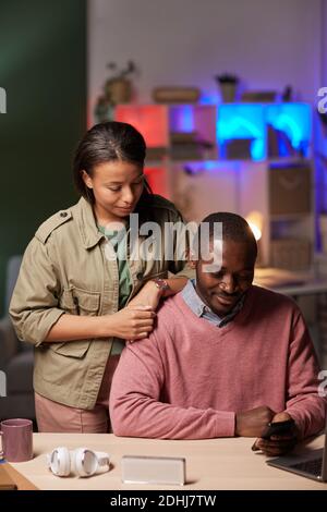 African man sitting at the table using mobile phone with his colleague standing near him they working till night Stock Photo
