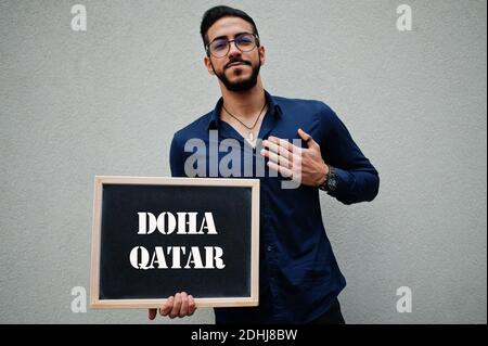 Arab man wear blue shirt and eyeglasses hold board with Doha Qatar inscription. Largest cities in islamic world concept. Stock Photo