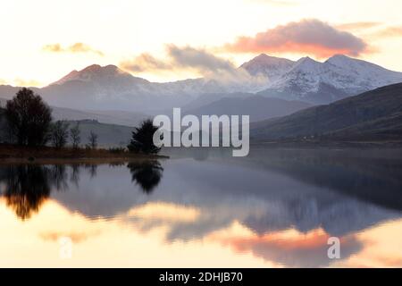 Sunset over the Snowdon horseshoe across showing recent snowfall beyond a flat calm Llynnau Mymbyr. Wales. Picture taken 5th December 2020