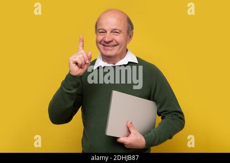 Smiling senior man holding alaptop pointing up, isolated on yellow background Stock Photo