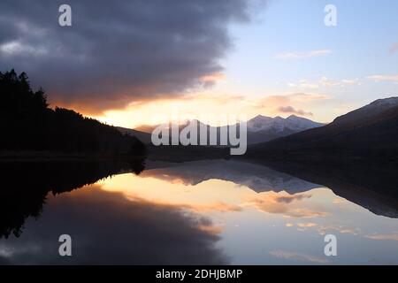 Sunset over the Snowdon horseshoe across showing recent snowfall beyond a flat calm Llynnau Mymbyr. Wales. Picture taken 5th December 2020