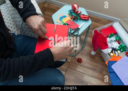 close up of person wrapping Christmas presents with paper, bows, sticky tape, labels in the home.  The man is sat down on an armchair cutting gift pap Stock Photo