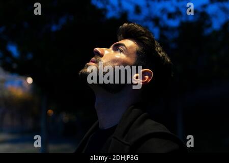Side view of bearded male contemplating and looking up while standing on dark street Stock Photo