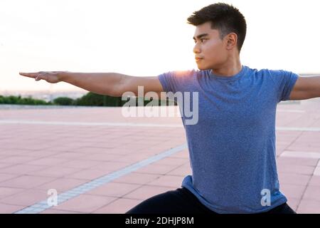 Focused Asian male standing on mat in urban park and doing yoga in Warrior pose while looking away Stock Photo