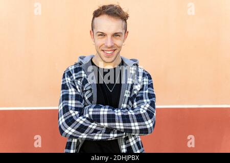 Positive confident young hipster guy in casual checkered shirt looking at camera and smiling while standing against orange wall Stock Photo