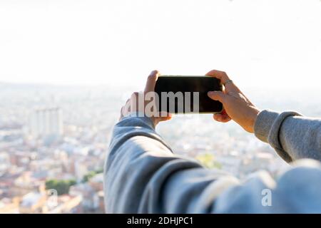 Cropped unrecognizable male traveler taking pictures with mobile phone while standing on green hill against blurred background in sunny day Stock Photo