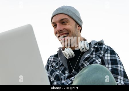 Low angle of happy young hipster male blogger in casual outfit with headphones on neck sitting on stone border and browsing laptop at sunset time Stock Photo