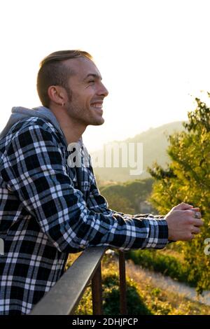 Side view of cheerful young hipster male traveler in casual plaid shirt leaning on railing and looking away while relaxing and contemplating nature in Stock Photo