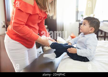 Side view of crop unrecognizable Woman putting on socks on legs of cute little boy sitting on bed at home Stock Photo