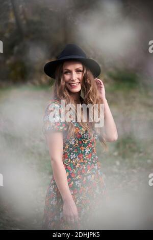 Side view of delighted female with long hair and in dress standing in woods while touching hat and looking at camera Stock Photo