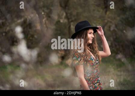 Side view of delighted female with long hair and in dress standing in woods while touching hat and looking away Stock Photo