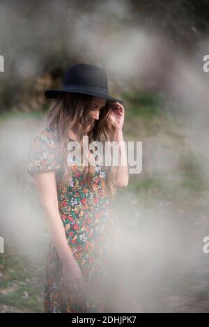Side view of delighted female with long hair and in dress standing in woods while touching hat and looking away Stock Photo
