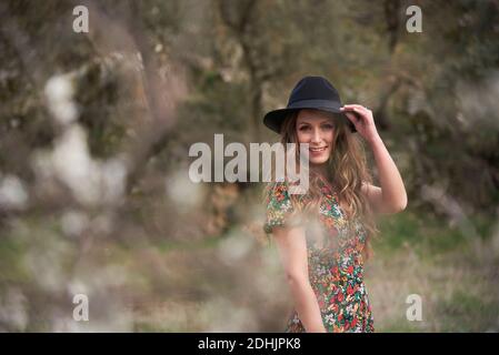 Side view of delighted female with long hair and in dress standing in woods while touching hat and looking at camera Stock Photo
