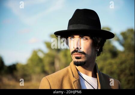 Young stylish hipster bearded male traveler with piercing wearing black hat looking away while standing on blurred countryside Stock Photo