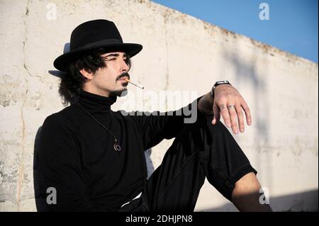 Side view of young bearded guy in stylish black clothes and hat smoking cigarette and looking away thoughtfully while sitting near shabby concrete wal Stock Photo