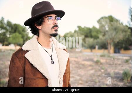 Handsome young bearded male in eyeglasses wearing trendy warm coat and hat looking away thoughtfully while standing against blurred trees in autumn da Stock Photo