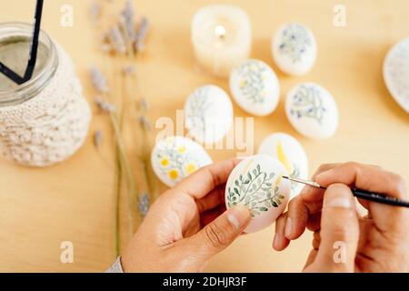 From above of crop unrecognizable person with paintbrush painting flowers on white eggs while preparing for Easter celebration at home Stock Photo
