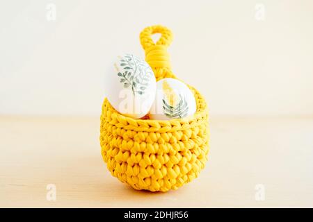 Handmade crochet basket with white chicken eggs painted with flowers placed on wooden table for Easter Stock Photo