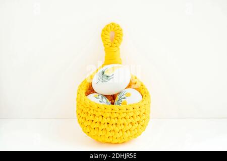 Handmade crochet basket with white chicken eggs painted with flowers placed on wooden table for Easter Stock Photo