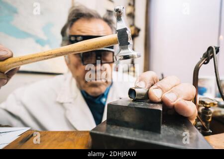 Focused skilled mature male master in glasses using hammer and chisel while working with gemstone in jewelry workshop Stock Photo