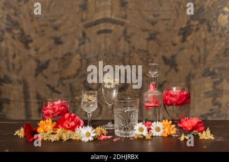 Beautiful stylish arrangement with various types of crystal glasses and bottle placed among assorted fresh flower buds on table against blurred orname Stock Photo