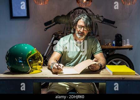 Front view of a man writing in a notebook on a wooden table next to a motorcycle helmet Stock Photo