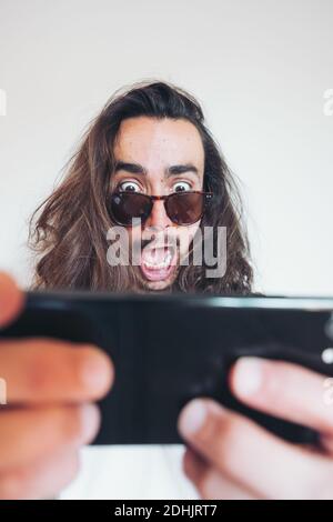 Overwhelmed young long haired hipster guy in sunglasses looking at screen of smartphone with astonished face expression while getting incredible news Stock Photo