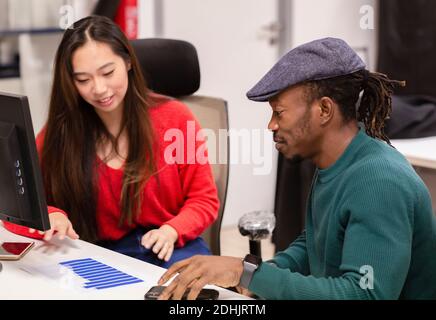 Positive young Asian Woman and African American male colleague in casual clothing sitting at table and analyzing business results while working with d Stock Photo