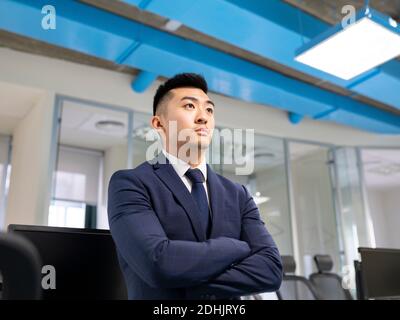 Serious young Asian male manager in formal suit looking away while standing near table with computers in contemporary coworking office Stock Photo