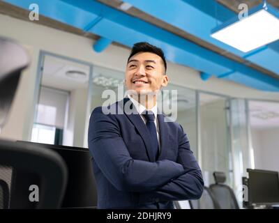 Cheerful young Asian male manager in formal suit looking away while standing near table with computers in contemporary coworking office Stock Photo