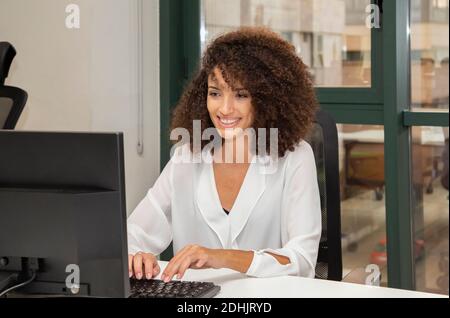 Young ethnic businessWoman with afro hairstyle sitting at table with computer while working on project in modern office Stock Photo