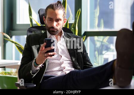 Concentrated adult bearded male entrepreneur in formal suit reading messages on mobile phone while sitting near window and having break during work in Stock Photo